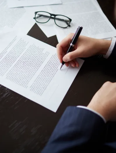 Mano de una persona firmando documentos junto a unas gafas sobre una mesa, relacionado con préstamos y créditos hipotecarios.